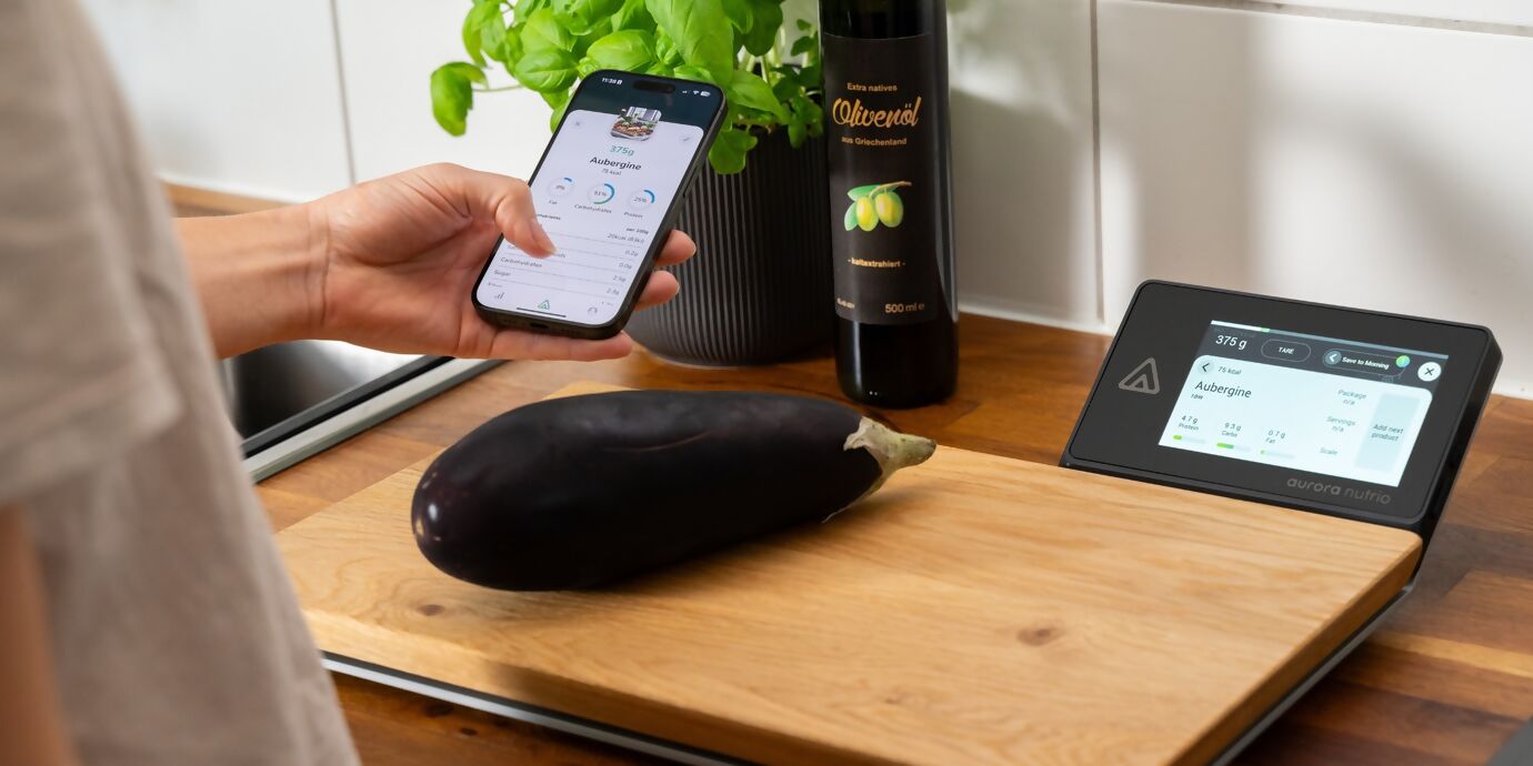 Aurora Nutrio Schneidebrett mit digitaler Anzeige und Aubergine auf dem Brett, das von einer Person mit Mobile Device in der Hand bedient wird, im Hintergrund Basilikum und Olivenöl
