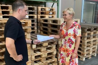 Mitarbeiterin übergibt Papiere an Mitarbeiterin aus der Logistik vor Palettenstapel