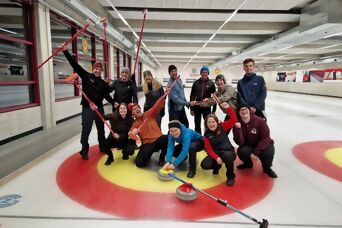 Ein Guppenfoto mit 12 Personen, die zusammen Curling gespielt hat jubelt mit Wischern und Curlingsteinen in die Kamera