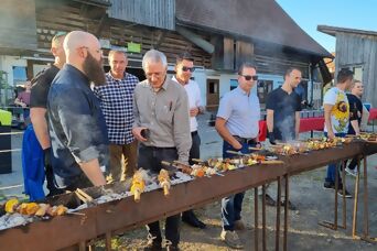 Eine Gruppe steht neben einem langen Grill auf dem Spiesse mit Gemüse und Fleisch zubereitet wird, bei schönem Wetter auf Bauernhof