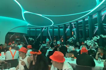 Viele Personen mit orangen Accessoires wie Hüten, Blumenketten, Hosenträgern und Fliegen sitzen im Restaurant