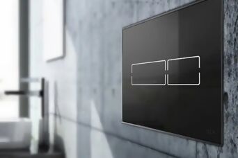 Black Tece Lux Mini toilet flush plate on a grey concrete wall in a bathroom