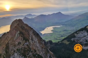 Luftaufnahme auf Gr.Mythen in Abendstimmung mit Blick ins Umland und Seen, rechts unten ein Plus-Icon auf orangem Kreis