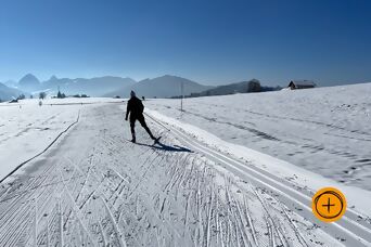 Langläufer auf Loipe im winterlichen Einsiedeln mit Blick auf Gr. Mythen bei blauem Himmel, rechts unten ein Plus-Icon auf orangem Kreis