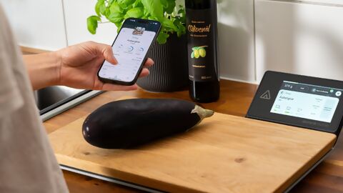 Aurora Nutrio Schneidebrett mit digitaler Anzeige und Aubergine auf dem Brett, das von einer Person mit Mobile Device in der Hand bedient wird, im Hintergrund Basilikum und Olivenöl