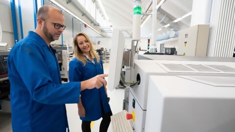 Zwei Mitarbeitende stehen in blauen ESD-Schutzmänteln vor Maschine in Elektronikproduktion und nehmen Einstellungen vor