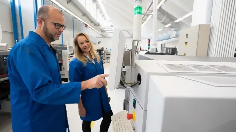 Zwei Mitarbeitende stehen in blauen ESD-Schutzmänteln vor Maschine in Elektronikproduktion und nehmen Einstellungen vor