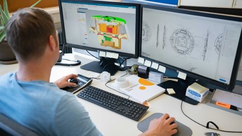 Mitarbeiter sitzt vor zwei Monitoren mit CAD-Zeichnung bei der Entwicklung einer mechanischen Komponente