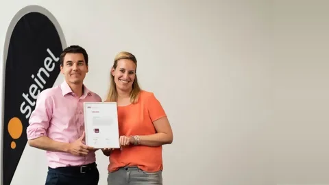 Marco Lang, CEO STEINEL Solutions und Melanie Niklaus, ehem. Head of Human Resources mit Urkunde von Fachstelle UND neben Steinel Beachflag
