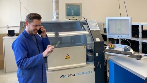 Mitarbeiter in blauem ESD-Mantel mit Mobiltelefon am Ohr und Leiterplatte in der Hand vor Selektrivlötmaschine